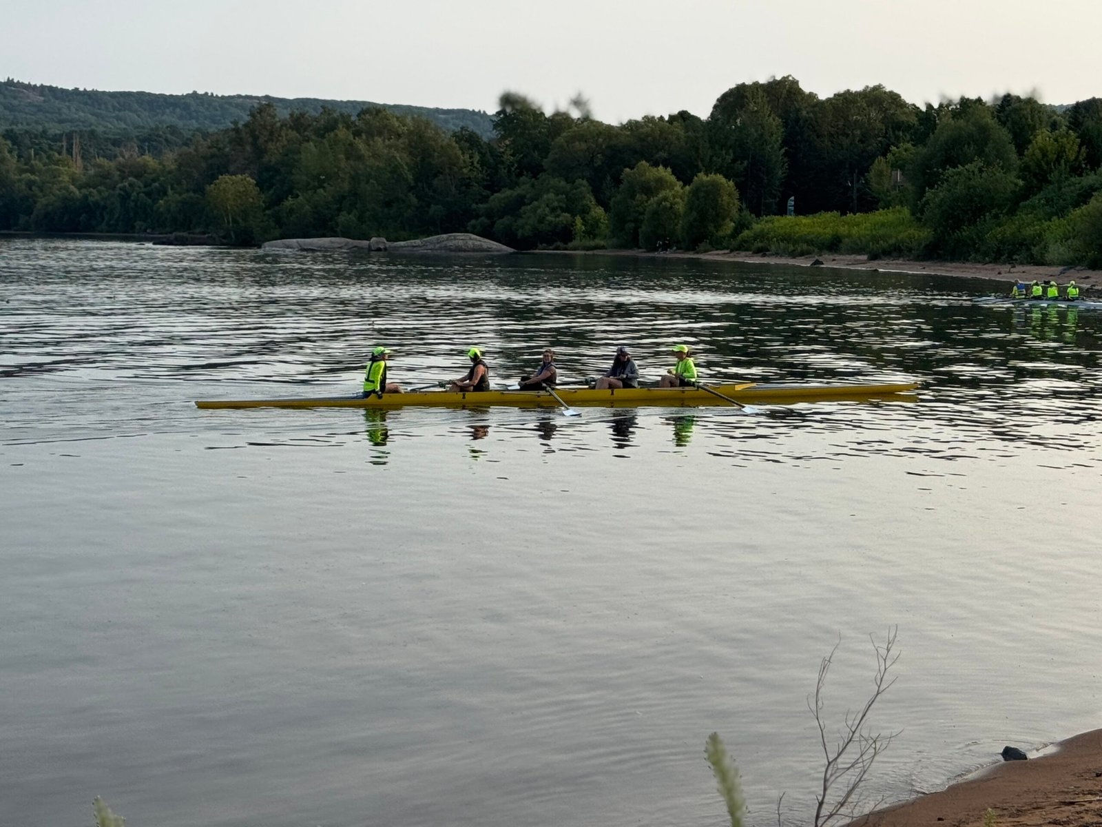 Four people in a yellow rowing boat from the UP rowing club paddle on a calm river near a forested shore, with another group of rowers visible in the distance. The peaceful scene is highlighted by water reflecting lush greenery.