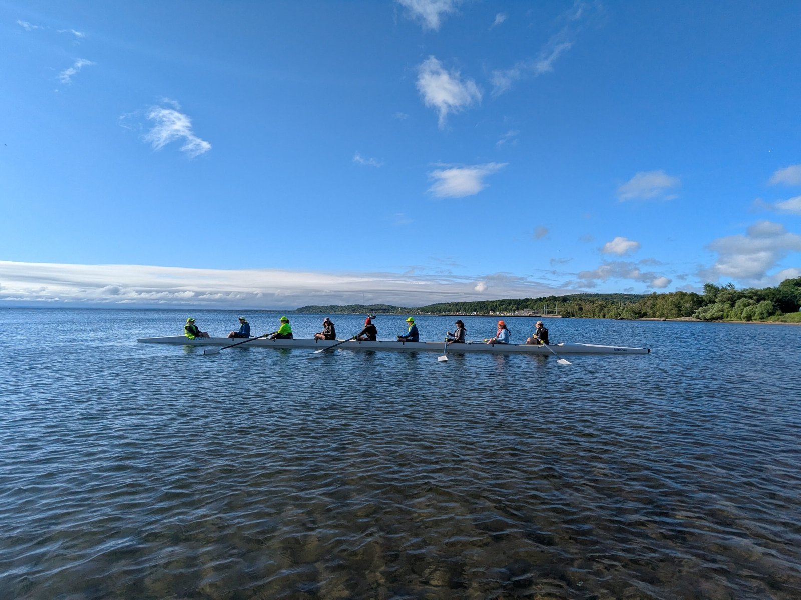 Eight people are rowing together in a long, narrow boat on a calm lake under a clear blue sky, with green trees and distant hills on the horizon.