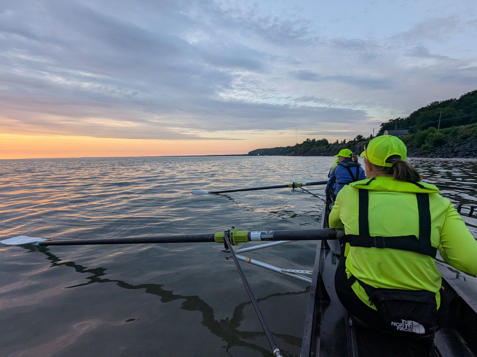 Rowers in bright yellow jackets and hats paddle on calm water at sunset, with a shoreline and trees visible on the right and a colorful sky in the background.