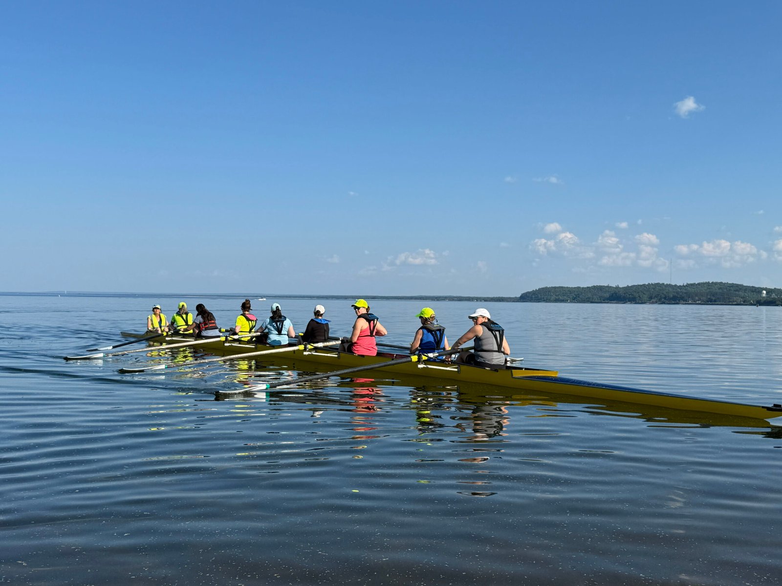 A group of people learn to row in Marquette, gliding together in two long, yellow boats on calm water under a clear blue sky, with distant green hills in the background.