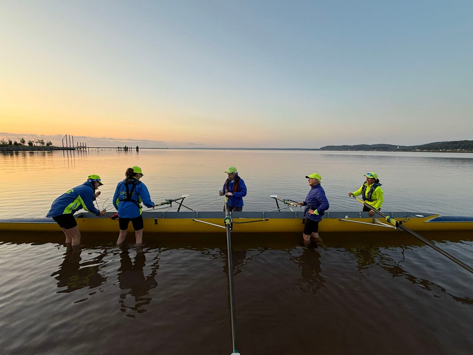 Five people in rowing gear stand in shallow water next to a yellow boat at sunrise or sunset, preparing for adult rowing for intermediate rowers on a calm lake with distant trees and hills in the background.