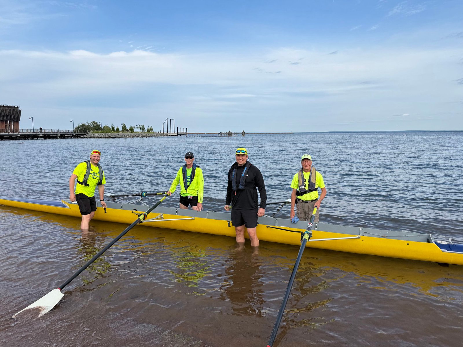 Four people in bright yellow shirts and life vests stand in shallow water beside a long yellow rowing boat, each holding oars—an ideal scene of adult rowing for intermediate rowers on a calm lake beneath a partly cloudy sky.