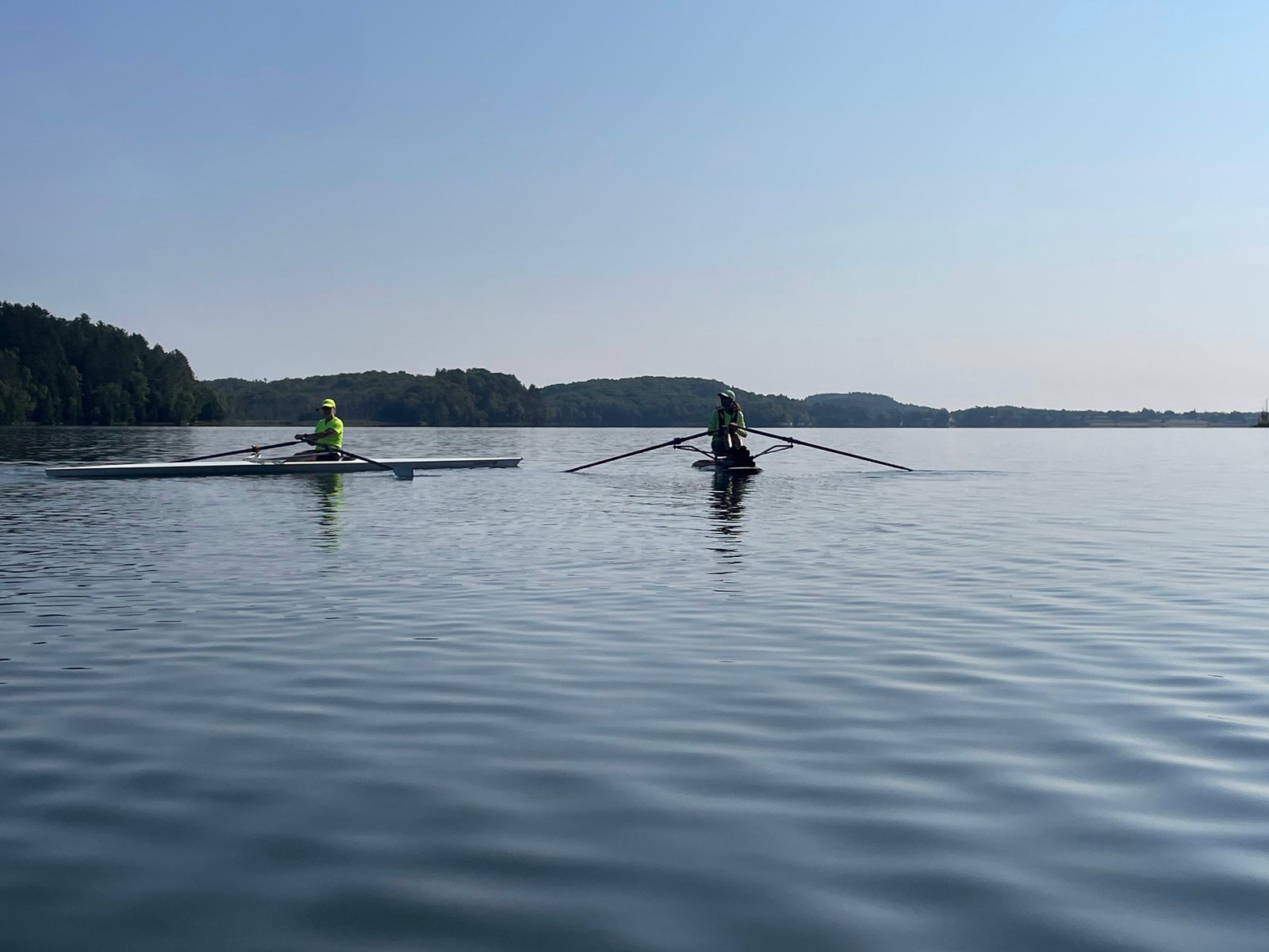 Two people, both adult rowing for intermediate rowers, glide small boats across a calm lake under a clear blue sky. Trees and hills line the distant shore, their reflections shimmering in the serene morning waters.