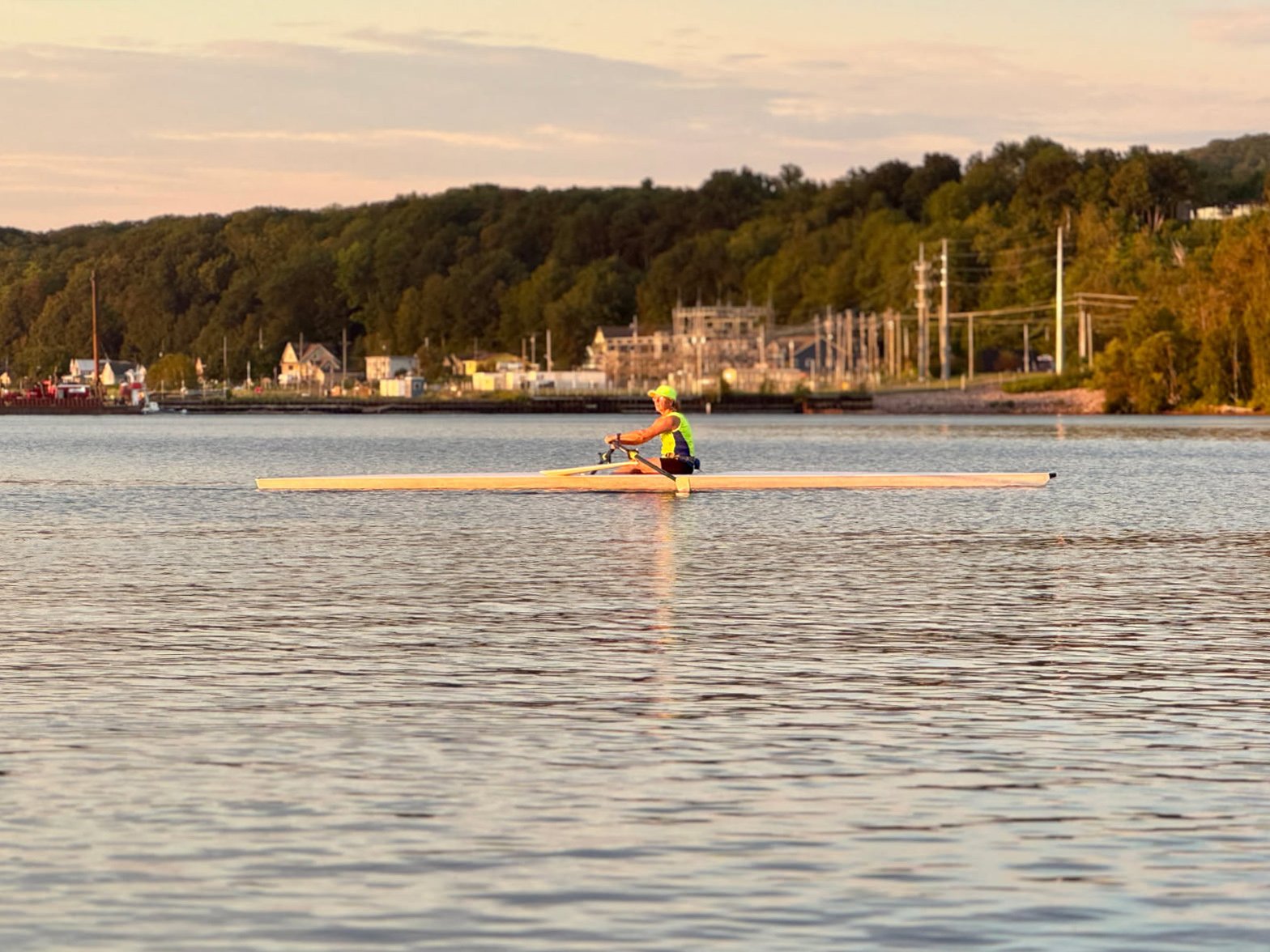 An adult rowing for intermediate rowers, wearing a neon vest and cap, rows a single scull boat on calm water, with a tree-lined shore and buildings in the background under a partly cloudy sky.